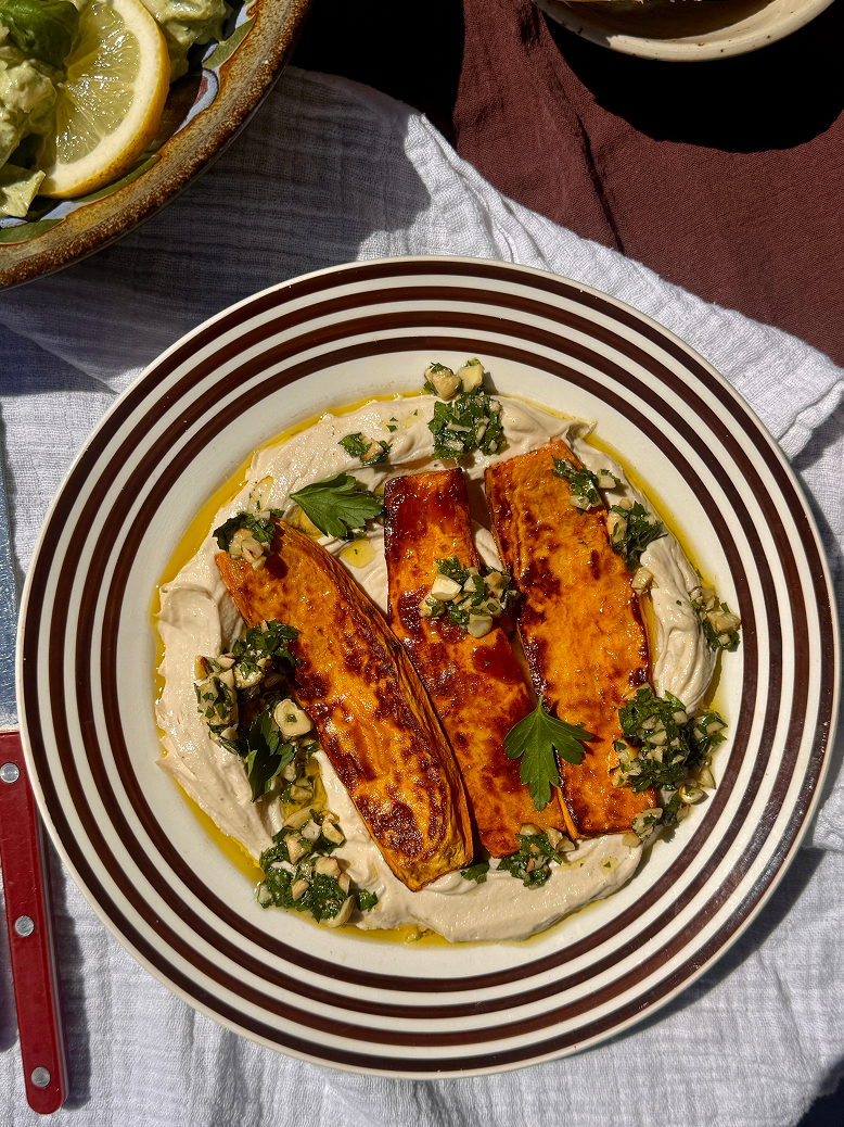 Tallerken med stegte søde kartofler, tahiniyoghurt og grøn topping på et dækket bord.